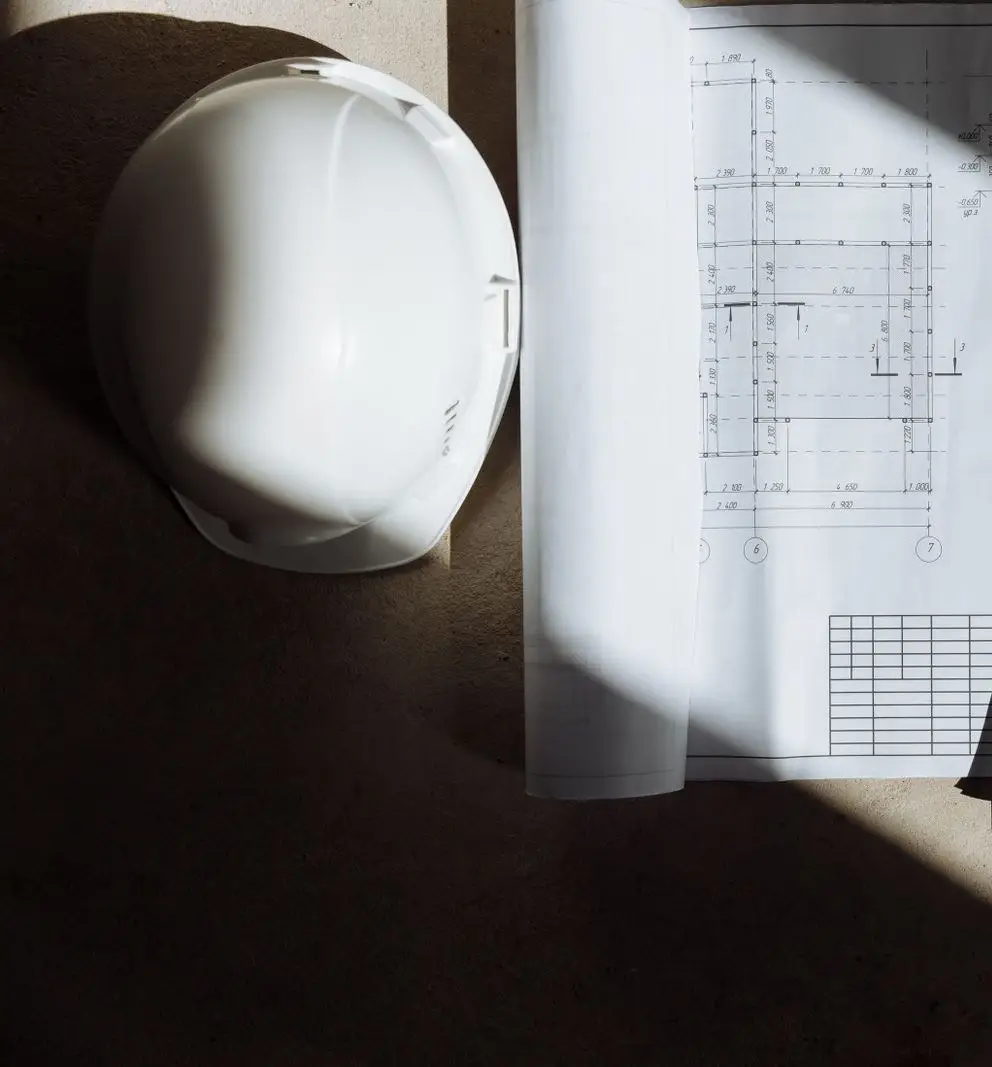 A white hard hat resting beside rolled structural drawings, lit by hard directional sunlight.