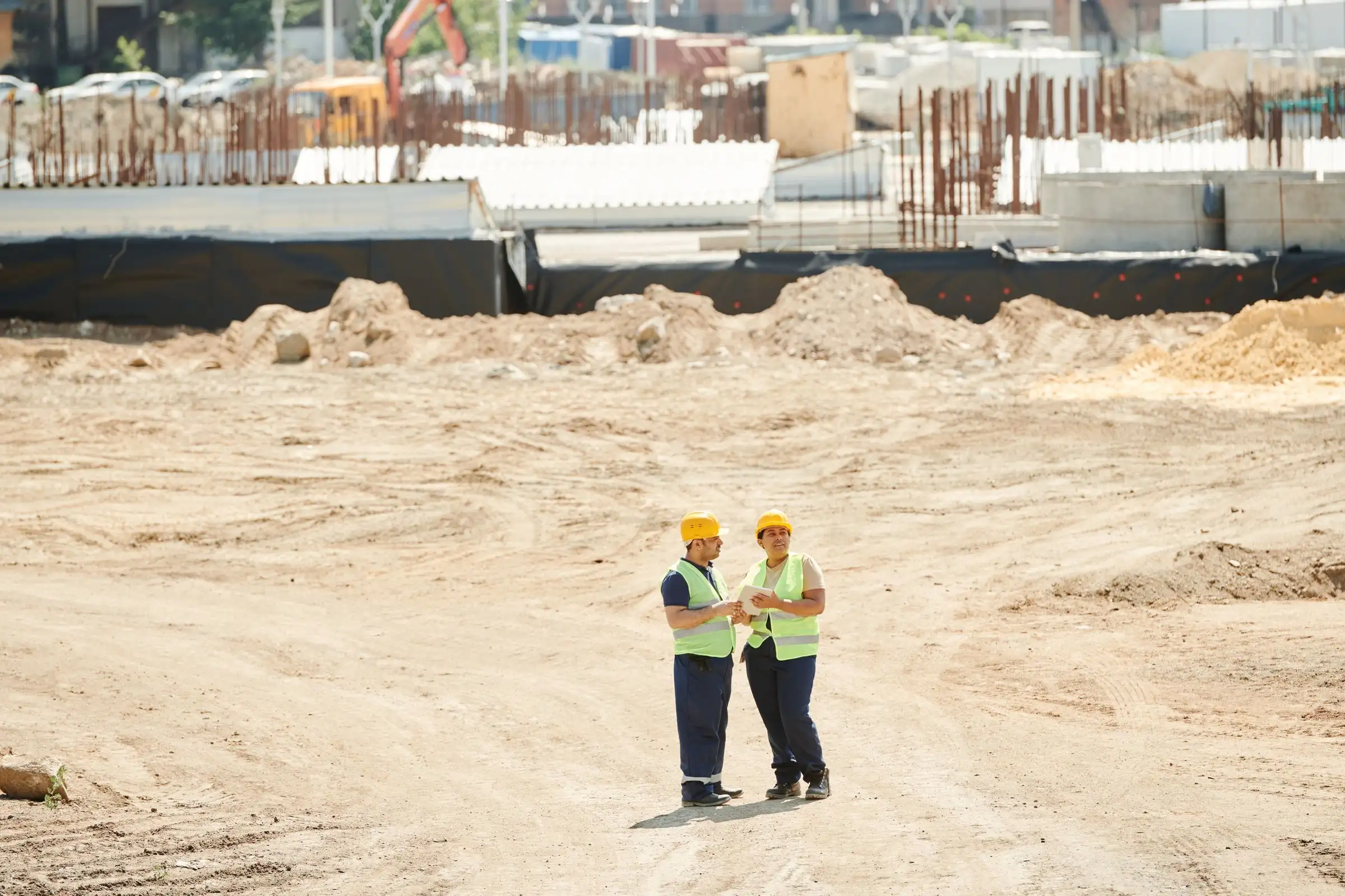 Two site staff in high-visibility vests reviewing work on the graded sand of an open construction site, with rebar starter bars and formwork behind them.