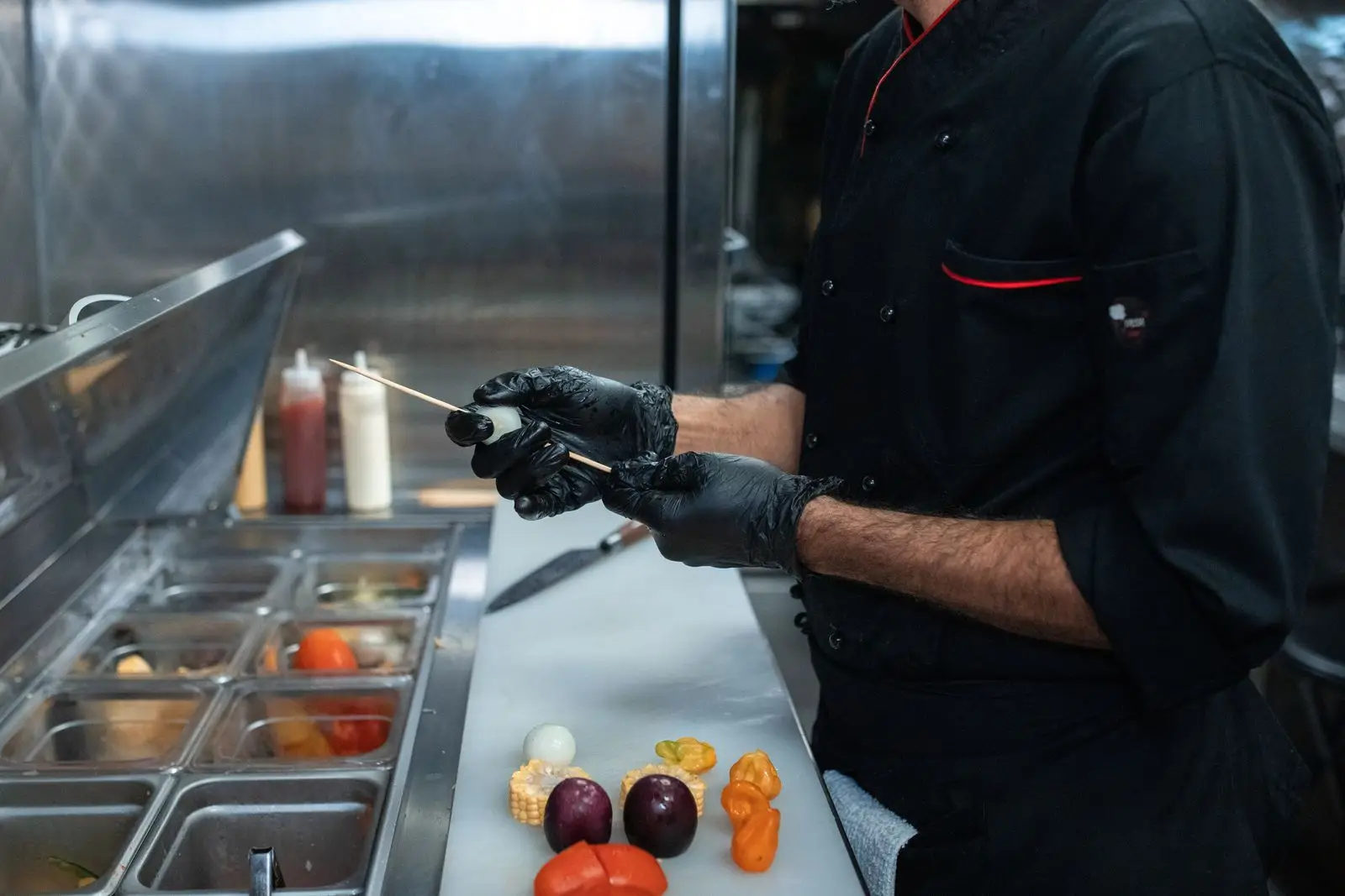 A kitchen worker in gloves preparing skewers at a stainless steel service counter in a commercial kitchen.