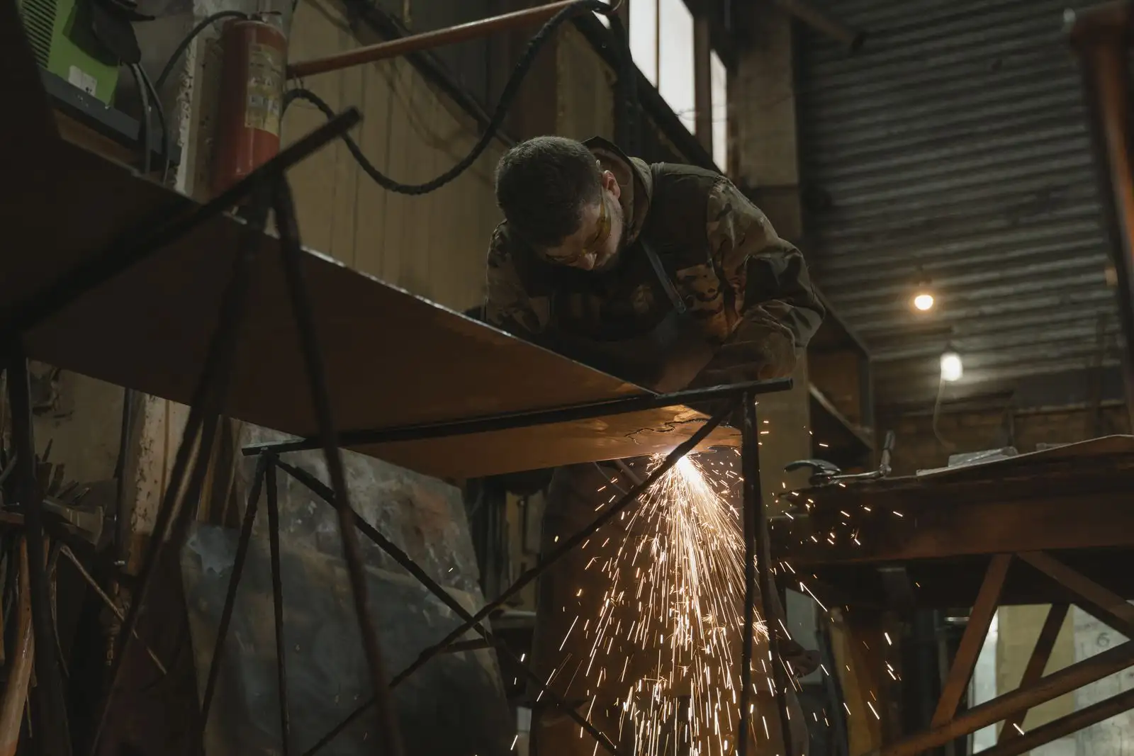 A welder cutting steel plate on trestles in a fabrication workshop, sparks falling below the work.