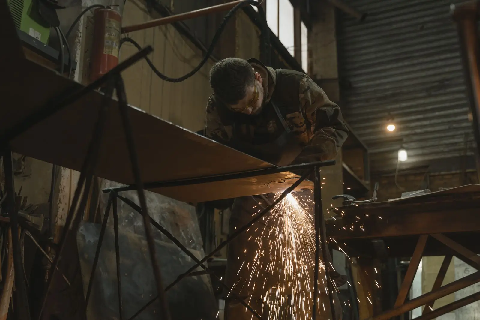 A welder cutting steel plate on trestles in a fabrication workshop, sparks falling below the work.