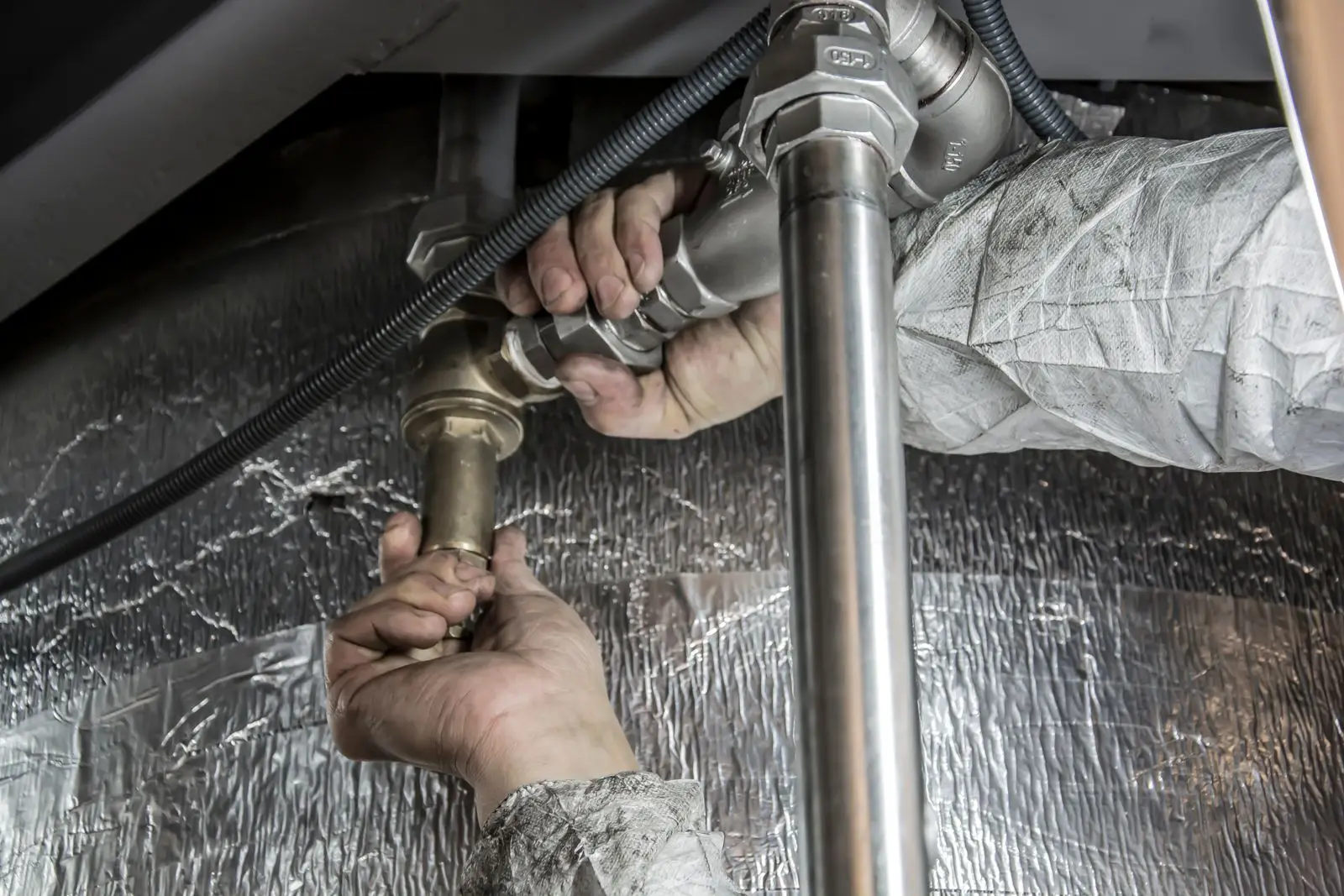 A plumber's hands tightening a threaded steel pipe union against insulated ductwork.